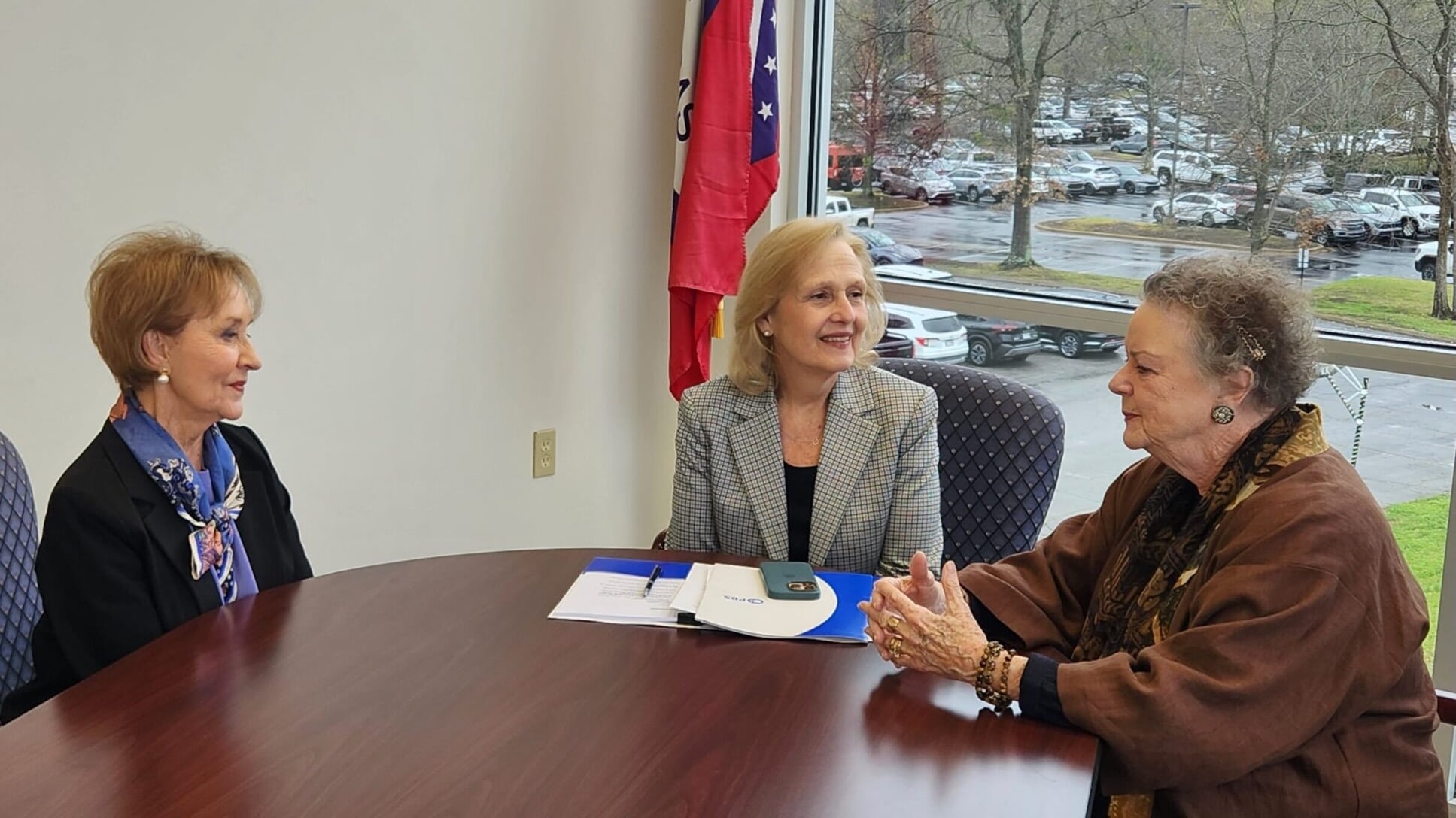 Former Arkansas First Lady Gay White, widow of Gov. Frank White; PBS CEO Paula Kerger; and former Arkansas First Lady Barbara Pryor, widow of Gov. David Pryor sit around a conference table in an office with large windows overlooking a parking lot on a rainy day. A folder and phone lie on the table, and an Arkansas state flag stands behind them.