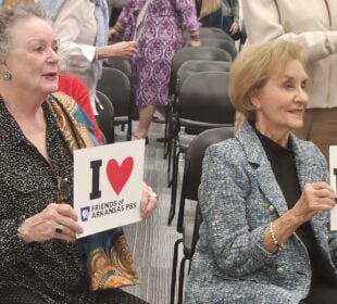 Barbara Pryor and Gay White are seated in a meeting room holding signs reading “I ♥ Friends of Arkansas PBS” and “I ♥ Arkansas PBS,” while other attendees stand behind them among rows of chairs.