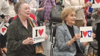 Barbara Pryor and Gay White are seated in a meeting room holding signs reading “I ♥ Friends of Arkansas PBS” and “I ♥ Arkansas PBS,” while other attendees stand behind them among rows of chairs.
