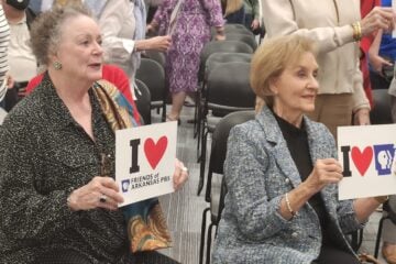 Barbara Pryor and Gay White are seated in a meeting room holding signs reading “I ♥ Friends of Arkansas PBS” and “I ♥ Arkansas PBS,” while other attendees stand behind them among rows of chairs.