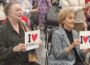 Barbara Pryor and Gay White are seated in a meeting room holding signs reading “I ♥ Friends of Arkansas PBS” and “I ♥ Arkansas PBS,” while other attendees stand behind them among rows of chairs.