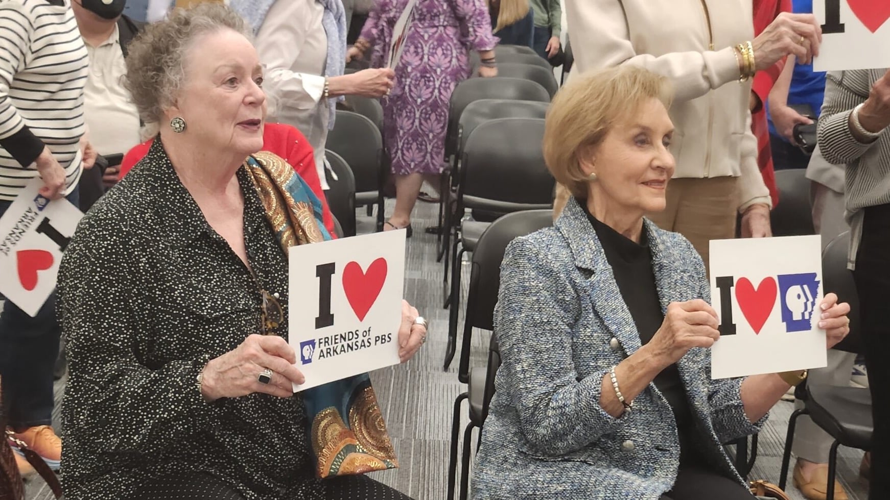 Barbara Pryor and Gay White are seated in a meeting room holding signs reading “I ♥ Friends of Arkansas PBS” and “I ♥ Arkansas PBS,” while other attendees stand behind them among rows of chairs.