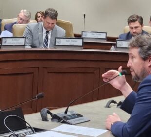 Little Rock Democratic Sen. Clarke Tucker speaks into a microphone during a legislative hearing, gesturing as several seated officials behind a curved dais review documents.