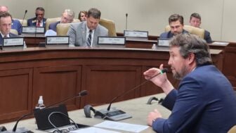Little Rock Democratic Sen. Clarke Tucker speaks into a microphone during a legislative hearing, gesturing as several seated officials behind a curved dais review documents.