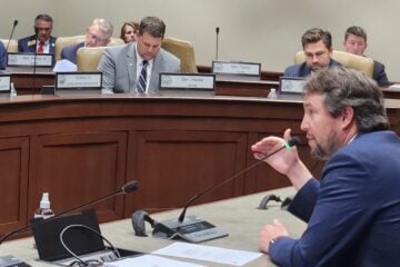 Little Rock Democratic Sen. Clarke Tucker speaks into a microphone during a legislative hearing, gesturing as several seated officials behind a curved dais review documents.