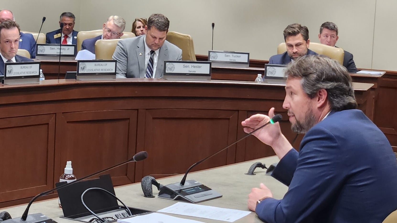 Little Rock Democratic Sen. Clarke Tucker speaks into a microphone during a legislative hearing, gesturing as several seated officials behind a curved dais review documents.
