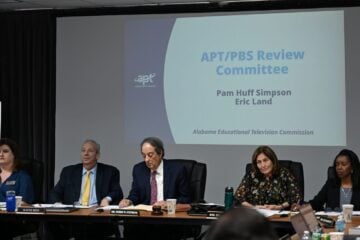 Members of the Alabama Educational Television Commission sit at a conference table during a meeting, with a projected slide behind them reading “APT/PBS Review Committee,” listing Pam Huff Simpson and Eric Land, Alabama Educational Television Commission.