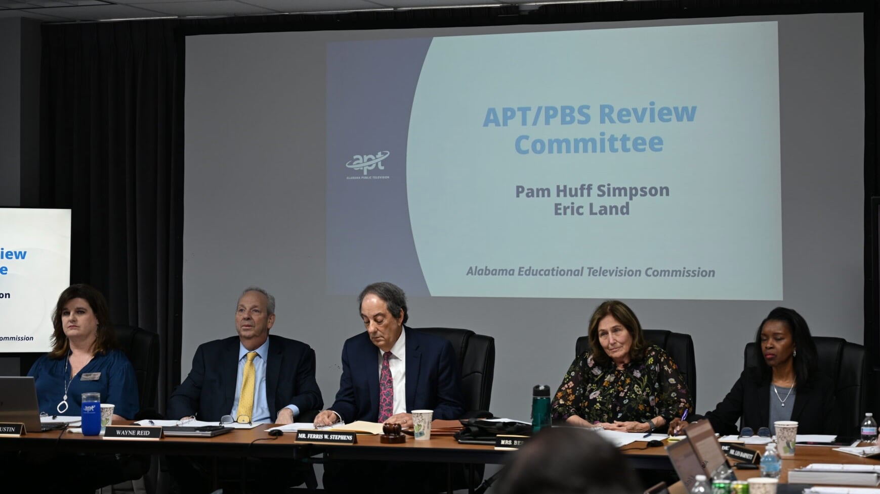 Members of the Alabama Educational Television Commission sit at a conference table during a meeting, with a projected slide behind them reading “APT/PBS Review Committee,” listing Pam Huff Simpson and Eric Land, Alabama Educational Television Commission.