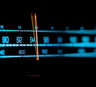 Close-up of an illuminated radio tuner dial showing FM and AM frequencies, with a glowing orange indicator centered around the mid-90s on the FM band against a dark background.