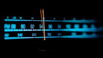 Close-up of an illuminated radio tuner dial showing FM and AM frequencies, with a glowing orange indicator centered around the mid-90s on the FM band against a dark background.