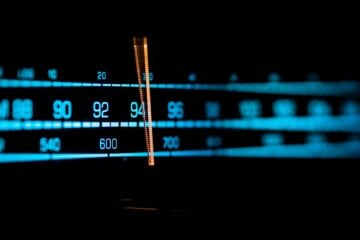 Close-up of an illuminated radio tuner dial showing FM and AM frequencies, with a glowing orange indicator centered around the mid-90s on the FM band against a dark background.