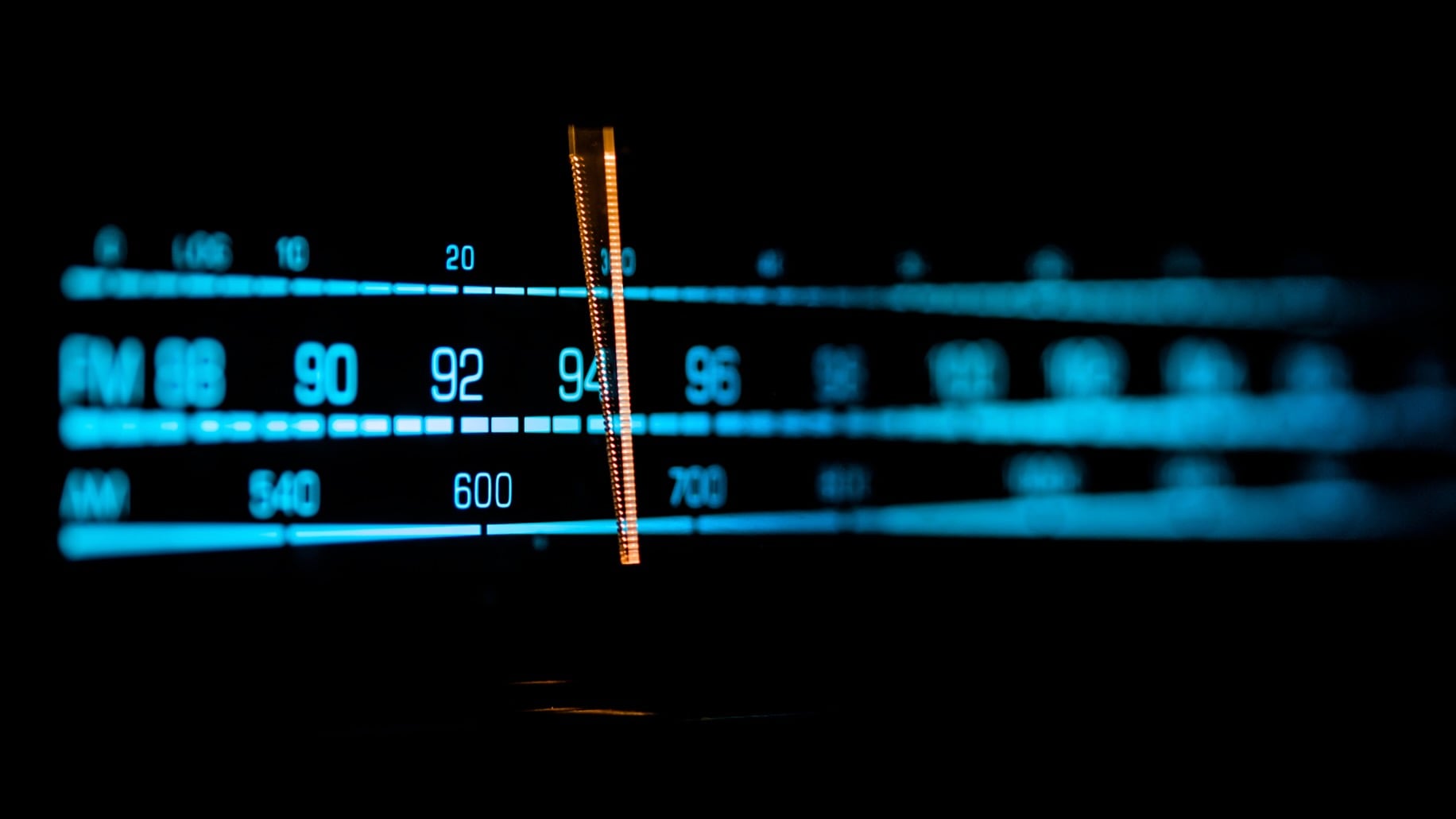 Close-up of an illuminated radio tuner dial showing FM and AM frequencies, with a glowing orange indicator centered around the mid-90s on the FM band against a dark background.