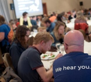 Attendees at the 2019 Third Coast International Audio Festival sit at round tables, eating and talking, with a person in the foreground wearing a shirt that reads “We hear you.”