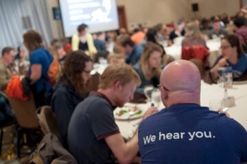 Attendees at the 2019 Third Coast International Audio Festival sit at round tables, eating and talking, with a person in the foreground wearing a shirt that reads “We hear you.”