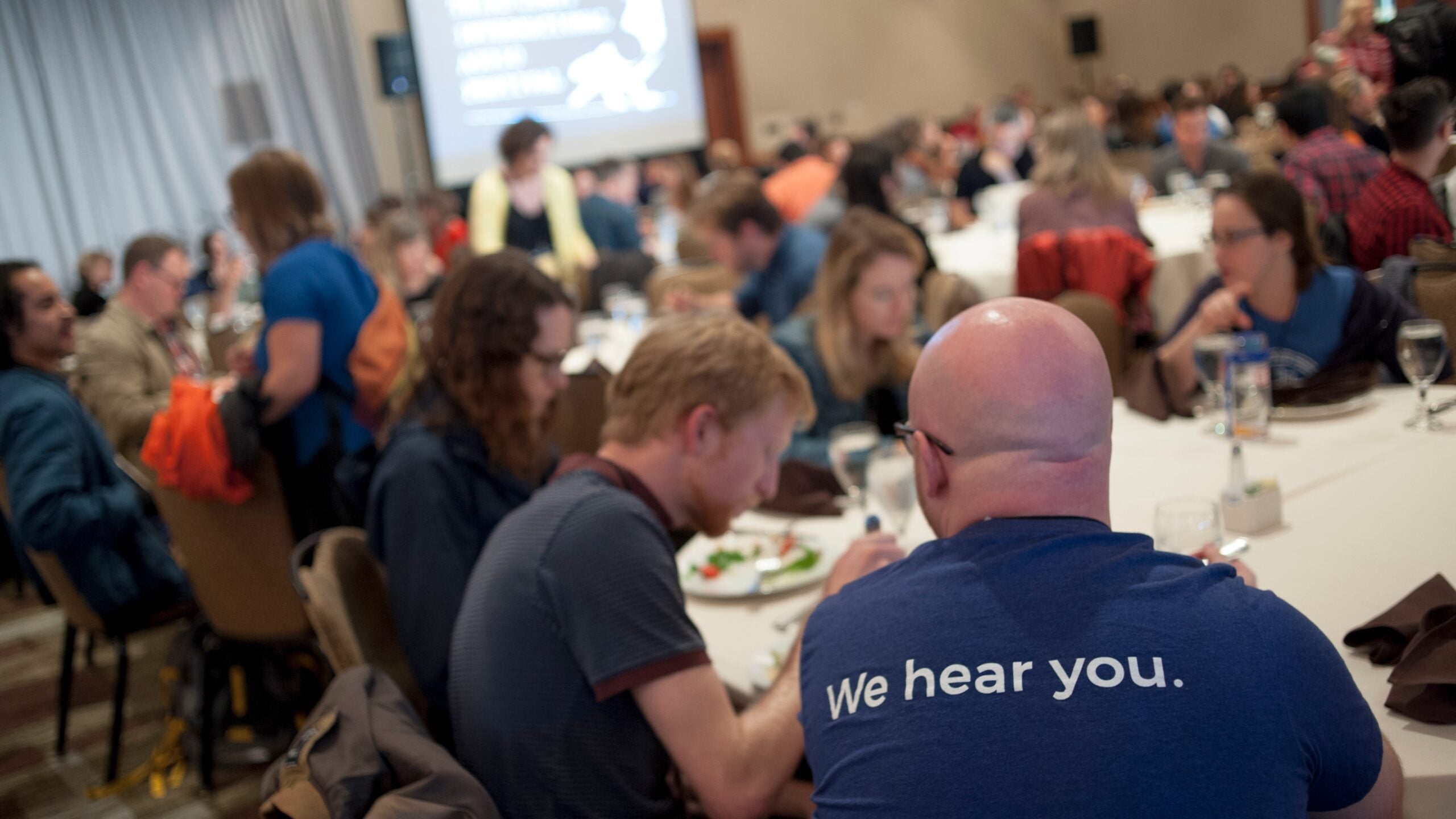 Attendees at the 2019 Third Coast International Audio Festival sit at round tables, eating and talking, with a person in the foreground wearing a shirt that reads “We hear you.”
