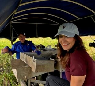 "LA64" host Karen LeBlanc smiles while sitting on a boat in a marsh as Barry Toups demonstrates crawfish harvesting equipment and Ryan Hamilton films the scene.