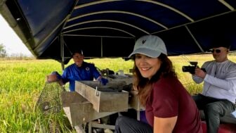 "LA64" host Karen LeBlanc smiles while sitting on a boat in a marsh as Barry Toups demonstrates crawfish harvesting equipment and Ryan Hamilton films the scene.