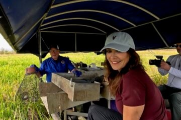 "LA64" host Karen LeBlanc smiles while sitting on a boat in a marsh as Barry Toups demonstrates crawfish harvesting equipment and Ryan Hamilton films the scene.