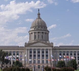 Front view of the Oklahoma State Capitol building in Oklahoma City, with its domed roof centered above rows of columns and state and tribal flags flying on the lawn under a partly cloudy sky.
