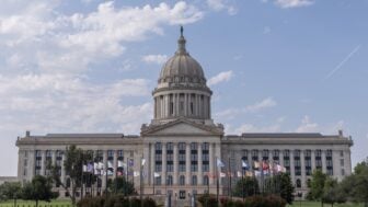 Front view of the Oklahoma State Capitol building in Oklahoma City, with its domed roof centered above rows of columns and state and tribal flags flying on the lawn under a partly cloudy sky.