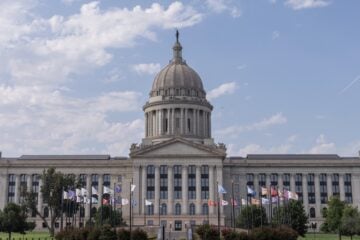 Front view of the Oklahoma State Capitol building in Oklahoma City, with its domed roof centered above rows of columns and state and tribal flags flying on the lawn under a partly cloudy sky.