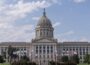 Front view of the Oklahoma State Capitol building in Oklahoma City, with its domed roof centered above rows of columns and state and tribal flags flying on the lawn under a partly cloudy sky.