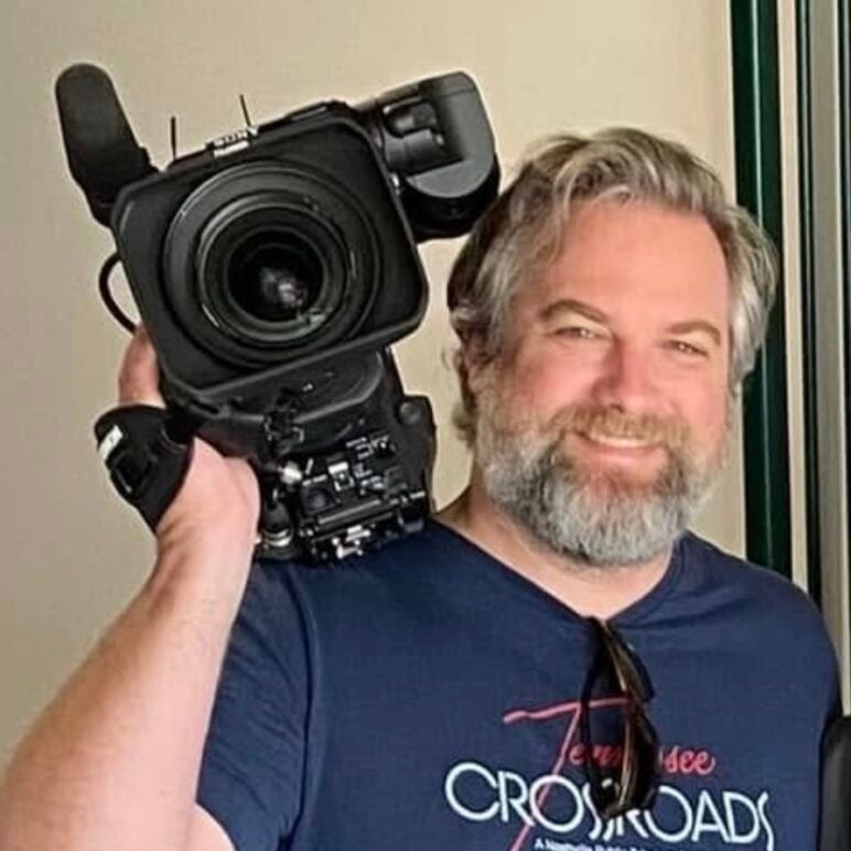 Paul Mojonnier smiles while holding a large professional video camera on his shoulder, wearing a blue T-shirt, in a close-up portrait.