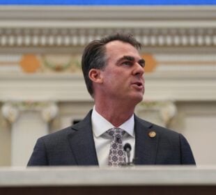 Oklahoma Governor Kevin Stitt, wearing a gray suit and patterned tie, speaks at a podium inside an ornate government chamber, viewed from a low angle with decorative architectural details blurred in the background.