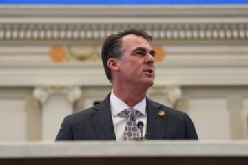 Oklahoma Governor Kevin Stitt, wearing a gray suit and patterned tie, speaks at a podium inside an ornate government chamber, viewed from a low angle with decorative architectural details blurred in the background.