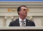 Oklahoma Governor Kevin Stitt, wearing a gray suit and patterned tie, speaks at a podium inside an ornate government chamber, viewed from a low angle with decorative architectural details blurred in the background.
