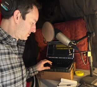 Wearing headphones, Ben Bradford listens to an interview on a laptop in his home recording studio lined with blankets and cushions and looks at notes on a notepad.