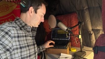 Wearing headphones, Ben Bradford listens to an interview on a laptop in his home recording studio lined with blankets and cushions and looks at notes on a notepad.