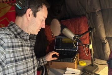 Wearing headphones, Ben Bradford listens to an interview on a laptop in his home recording studio lined with blankets and cushions and looks at notes on a notepad.