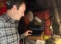 Wearing headphones, Ben Bradford listens to an interview on a laptop in his home recording studio lined with blankets and cushions and looks at notes on a notepad.