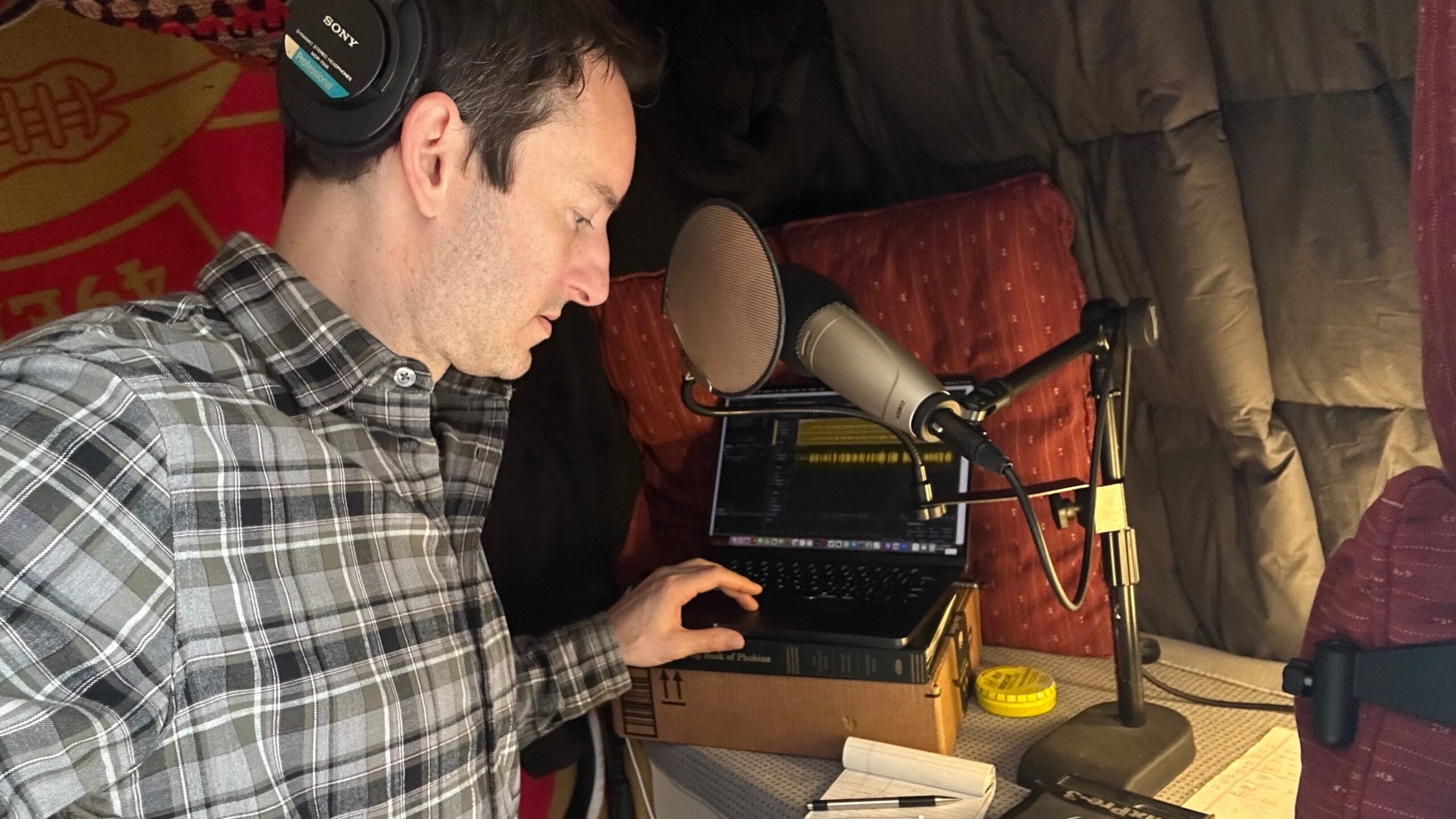 Wearing headphones, Ben Bradford listens to an interview on a laptop in his home recording studio lined with blankets and cushions and looks at notes on a notepad.