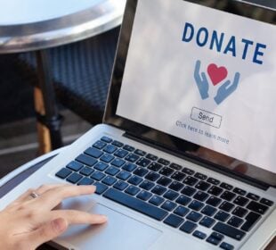 A person uses a laptop displaying an online donation page with a heart icon and a donation button, illustrating digital charitable giving.