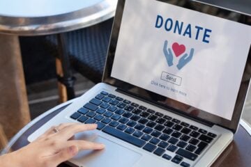 A person uses a laptop displaying an online donation page with a heart icon and a donation button, illustrating digital charitable giving.