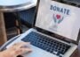 A person uses a laptop displaying an online donation page with a heart icon and a donation button, illustrating digital charitable giving.