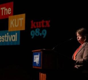 KUT GM Debbie Hiott stands at a podium on a dark stage during The KUT News Festival. Large projected logos reading “The KUT News Festival” and “kutx 98.9” appear behind her. Hiott, wearing a blazer and orange top, is illuminated by stage lighting as she addresses the audience.