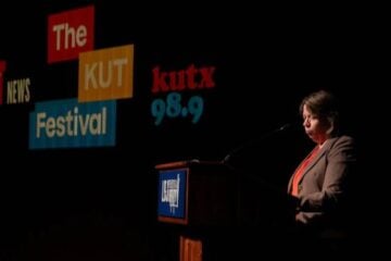 KUT GM Debbie Hiott stands at a podium on a dark stage during The KUT News Festival. Large projected logos reading “The KUT News Festival” and “kutx 98.9” appear behind her. Hiott, wearing a blazer and orange top, is illuminated by stage lighting as she addresses the audience.