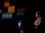 KUT GM Debbie Hiott stands at a podium on a dark stage during The KUT News Festival. Large projected logos reading “The KUT News Festival” and “kutx 98.9” appear behind her. Hiott, wearing a blazer and orange top, is illuminated by stage lighting as she addresses the audience.