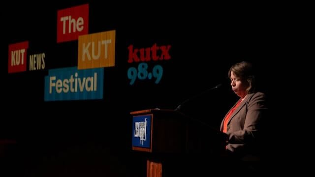 KUT GM Debbie Hiott stands at a podium on a dark stage during The KUT News Festival. Large projected logos reading “The KUT News Festival” and “kutx 98.9” appear behind her. Hiott, wearing a blazer and orange top, is illuminated by stage lighting as she addresses the audience.