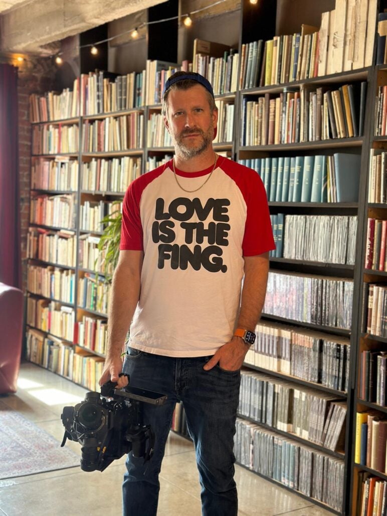 Adam Mason stands in front of floor-to-ceiling bookshelves filled with books and records, holding a professional video camera. He wears a white-and-red baseball-style shirt with bold lettering that says "LOVE IS THE FING.", jeans and a backward cap in a warmly lit room with exposed brick and string lights.