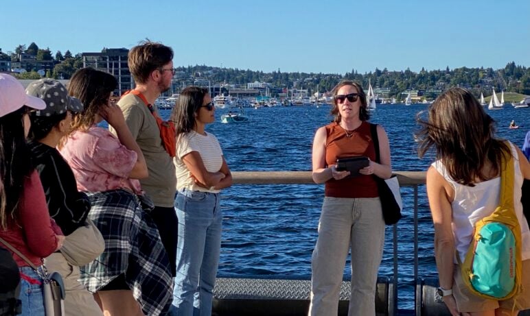 A group of people stands along a waterfront listening to Cara Kuhlman speak during an outdoor walking tour. Sailboats dot the water behind them, with a shoreline and buildings visible across the bay on a sunny day.
