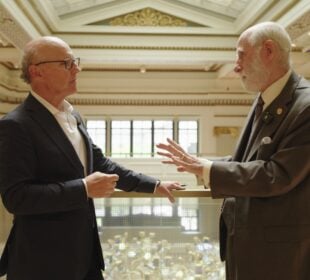 David McCourt, left, and internet pioneer Vint Cerf stand on a balcony inside a grand hall, facing one another as they talk. Cerf gestures with one hand while McCourt listens, with the ornate interior visible in the background.