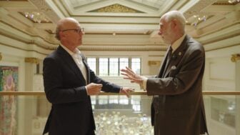 David McCourt, left, and internet pioneer Vint Cerf stand on a balcony inside a grand hall, facing one another as they talk. Cerf gestures with one hand while McCourt listens, with the ornate interior visible in the background.