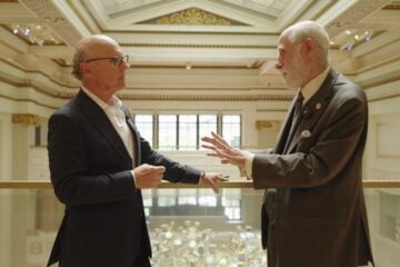 David McCourt, left, and internet pioneer Vint Cerf stand on a balcony inside a grand hall, facing one another as they talk. Cerf gestures with one hand while McCourt listens, with the ornate interior visible in the background.
