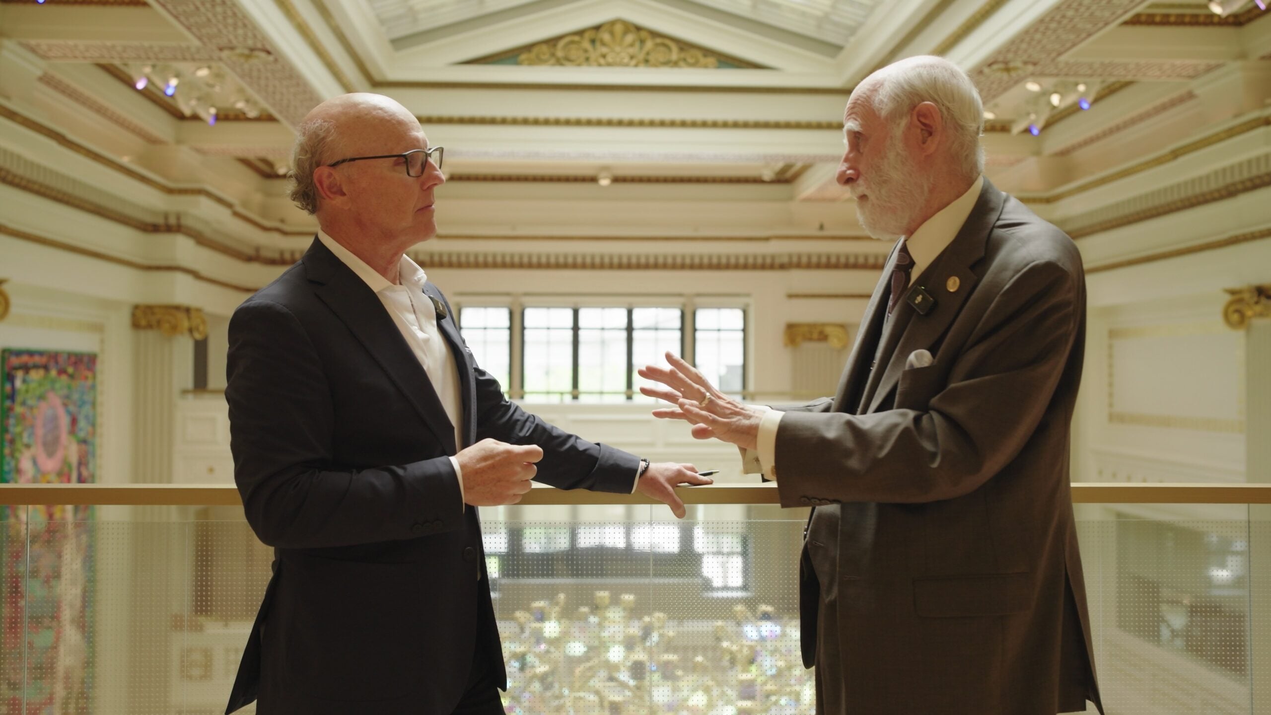 David McCourt, left, and internet pioneer Vint Cerf stand on a balcony inside a grand hall, facing one another as they talk. Cerf gestures with one hand while McCourt listens, with the ornate interior visible in the background.