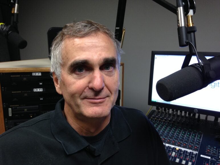 Karl Gehrke sits in a radio studio beside a suspended broadcast microphone. Audio equipment, a mixing console and computer monitors are visible behind him as he looks toward the camera.