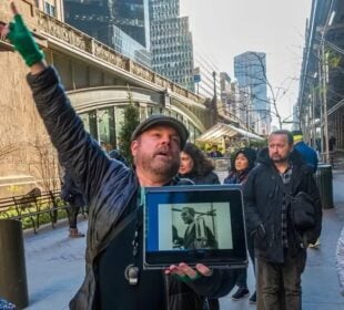 Justin Rivers, chief experience officer at Untapped New York, stands on a city sidewalk speaking to a group of participants while holding a tablet displaying a historic photograph. Tall buildings, scaffolding and pedestrians line the street in the background.